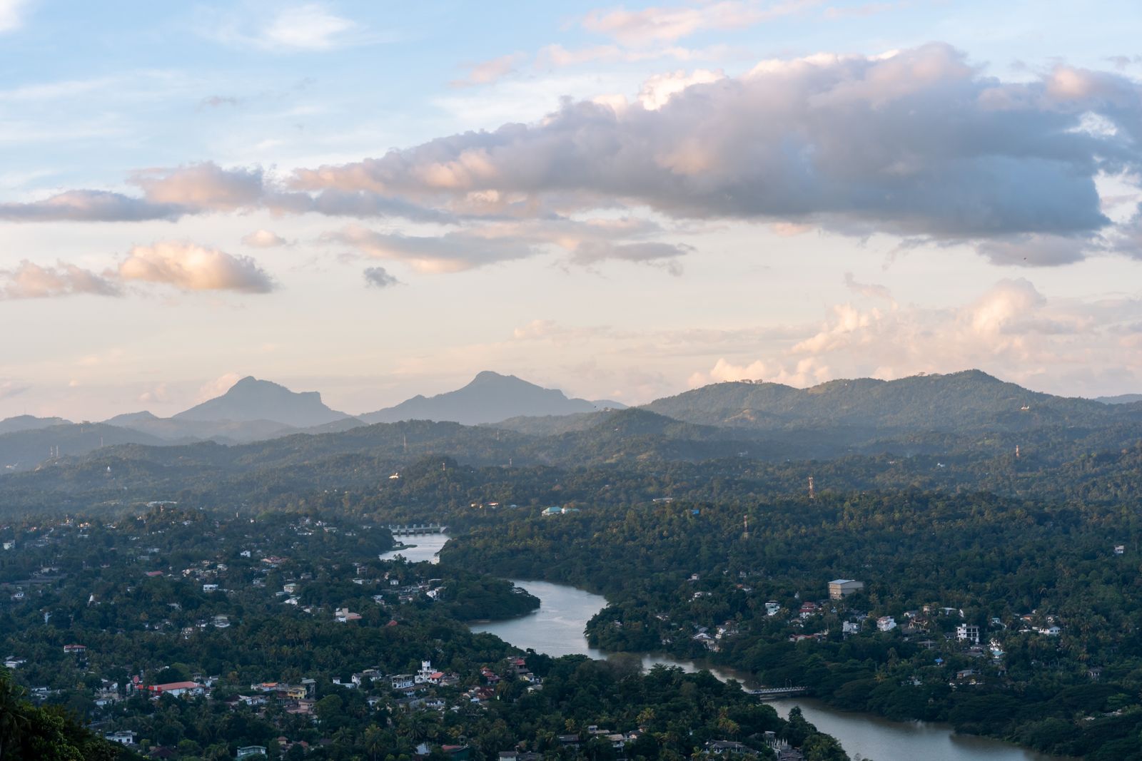 Aerial view of lush green Sri Lankan hills with a calm river and distant mountains under a cloudy sky.